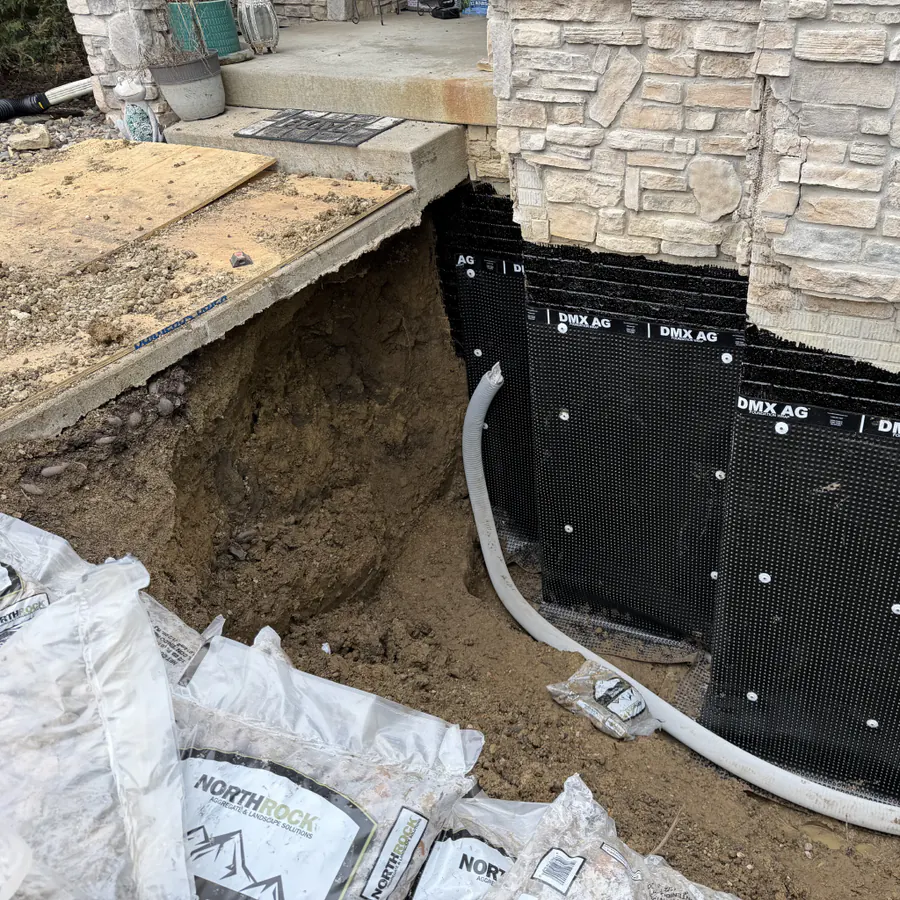 Exterior foundation repair and waterproofing in Iowa: excavation trench along a stone-veneer wall with DMX AG membrane, white perimeter drainage pipe in the trench, and aggregate bags ready for backfill